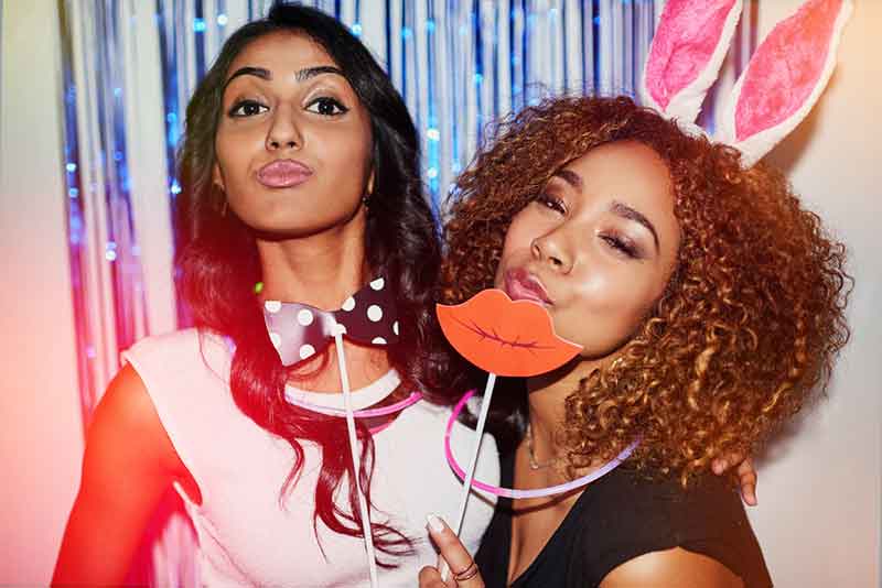 Two friends in playful props at a pink-lit photo booth.